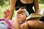 Young girl playing with blade of grass resting head on mother lap\n