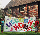 Porträt von zwei kleinen Kindern, die ein buntes Willkommen-Zuhause-Schild halten