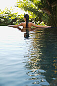 Back of young woman standing in an exotic swimming pool with arms stretched\n