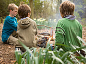 Three boys sitting around campfire cooking fish\n