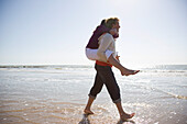 Mann trägt Frau auf dem Rücken und geht am Strand spazieren