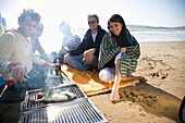 Friends sitting around a barbeque on the beach\n