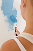 Back view of woman holding paint brush and painting a white wall with blue paint\n