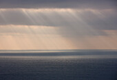 Grey sky with rain and sun rays shining on the ocean\n