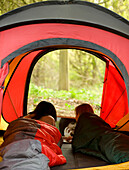 Back view of two teenaged girls lying in a tent looking at a photo on a laptop computer\n