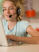 Lächelndes junges Mädchen mit Kopfhörern und Mikrofon sitzt am Schreibtisch und tippt auf einem Laptop Computer