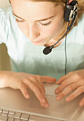 Close up of smiling young girl wearing earphones and microphone typing on a laptop computer\n