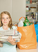 Junges Mädchen steht neben Recyclingsäcken und hält einen Stapel Zeitungen