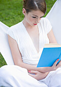 Teenaged girl sitting on high back chair reading a book\n