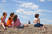 Kinder sitzen am Strand und beobachten einen Jungen, der einen Drachen steigen lässt