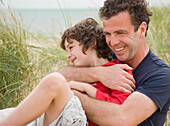 Father and son sitting on a beach smiling and embracing\n