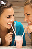 Two smiling teenaged girls drinking milk-shake with a straw from the same glass\n
