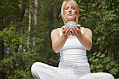 Junge Frau in Meditation im Schneidersitz vor einem Wald, eine Kugel in den Händen haltend