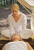 Female massage therapist sitting behind a woman massaging her scalp under a tent\n