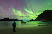 Mann mit Rucksack bewundert die hellen grünen Lichter der Aurora Borealis (Nordlichter) vom Haukland Strand, Lofoten Inseln, Nordland, Norwegen, Skandinavien, Europa