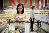 Frau beim Einkaufen im Supermarkt und mit Handy, um Preise zu vergleichen oder die Einkaufsliste zu überprüfen