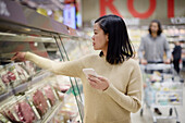 Frau kauft im Supermarkt ein und benutzt ihr Handy, um Preise zu vergleichen oder die Einkaufsliste zu prüfen