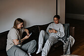 Female couple sitting together on sofa and using cell phones\n