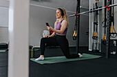 Young woman using phone while stretching on mat in gym\n