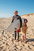 Blondes Mädchen mit Vater trägt Surfbrett am Strand