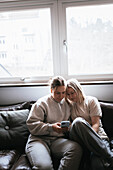Homosexual couple sitting on sofa and using cell phone\n
