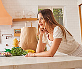 Smiling woman with paper shopping bag and groceries in kitchen\n