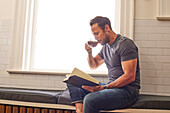 Mann liest Buch und trinkt Tee am Fenster zu Hause