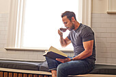 Man reading book and drinking tea by window at home\n