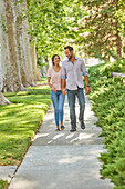 Lächelndes Paar hält sich an den Händen und geht auf einem mit Bäumen gesäumten Gehweg spazieren