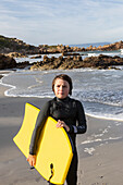South Africa, Hermanus, Portrait of boy (10-11) standing with body board by Atlantic Ocean in Voelklip Beach\n