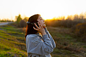 Side view of woman listening to music with closed eyes in meadow at sunset\n