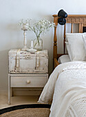 Wooden bedside table with vintage suitcase and vase with gypsophila next to a rustic bed