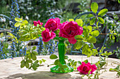 Rote Rosenblüten (Rosa) dekoriert mit Stein auf Stielvase und Zweigen auf Gartentisch