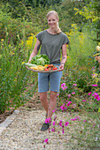 Junge Frau trägt Tablett aus Korb mit frisch geerntetem Gemüse, Eisbergsalat, Zucchini, Tomaten und Bohnen 'Neckargold' (Phaseolus) vor Blumenbeet