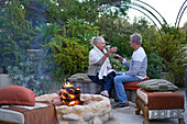 Senior couple drinking wine by fire pit on garden patio