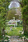 Table set in the garden for Easter breakfast with Easter nest and coloured eggs, bouquet of flowers, dog in the meadow, archway through climbing plants