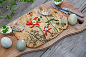 Easter flatbread decorated with herbs, fried onions and blossom of pepper strips on wooden board, Easter eggs, cutlery and daisies
