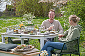 Young couple sitting at a set table for Easter breakfast with Easter nest, colored eggs, cereal bowls and water jug, and bouquet of flowers in the garden