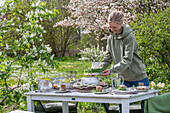 Young woman places bouquet of flowers on table set for Easter breakfast with Easter nest and colored eggs, in front of flowering bushes