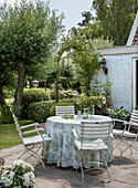 Runder Gartentisch mit weiß-grüner Tischdecke und Gartenstühlen im Sommergarten