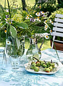 Glasvase und Karaffe mit Wiesenblumen und Teller mit Äpfeln auf einem gedeckten Gartentisch