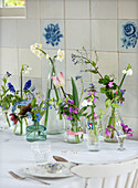 Spring flowers in various glass vases on a white table in front of a tiled backdrop with a floral pattern