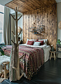 Bed with patterned bedspread in bedroom with wooden wall paneling and clothes rack made of natural wood