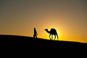 Marokko,Silhouette von Berber 'Blauer Mann' führt Kamel über Sanddünen in der Abenddämmerung im Erg Chebbi-Gebiet,Sahara-Wüste bei Merzouga