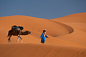 Morocco,Berber 'Blue man' leading camel across sand dunes in Erg Chebbi area,Sahara Desert near Merzouga