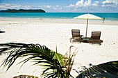 Malaysia,Pulau Langkawi,Liegestühle unter Sonnenschirm am weißen Sandstrand mit Palmen mit Blick auf das blaue Meer,Pantai Cenang (Cenang Strand)