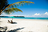 Malaysia,Pulau Langkawi,Deck chairs under parasol on white sandy beach with palm trees overlooking blue sea,Pantai Cenang (Cenang beach)