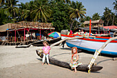 Silvie Reynolds, 6 Jahre, und Isla Reynolds, 4 Jahre, stehen vor den vielen traditionellen Booten und Strandrestaurants, die den Strand von Palolem, Goa, Indien, säumen.