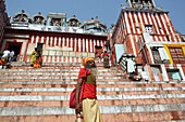 Hindu-Sadhu, der an einem Hindu-Tempel und den farbenfrohen Stufen des Bade-Ghat oberhalb des Ganges vorbeigeht. Die Kultur von Varanasi ist eng mit dem Fluss Ganges und seiner religiösen Bedeutung verbunden: Sie ist die "religiöse Hauptstadt Indiens" und ein wichtiges Zentrum für die