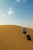 Barfüßiger Mann mit Koffer geht Sanddüne hinauf,Dubai,Vereinigte Arabische Emirate